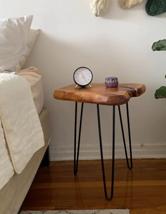 A small wooden side table with black metal legs placed next to a bed, topped with a round clock and a colorful candle.