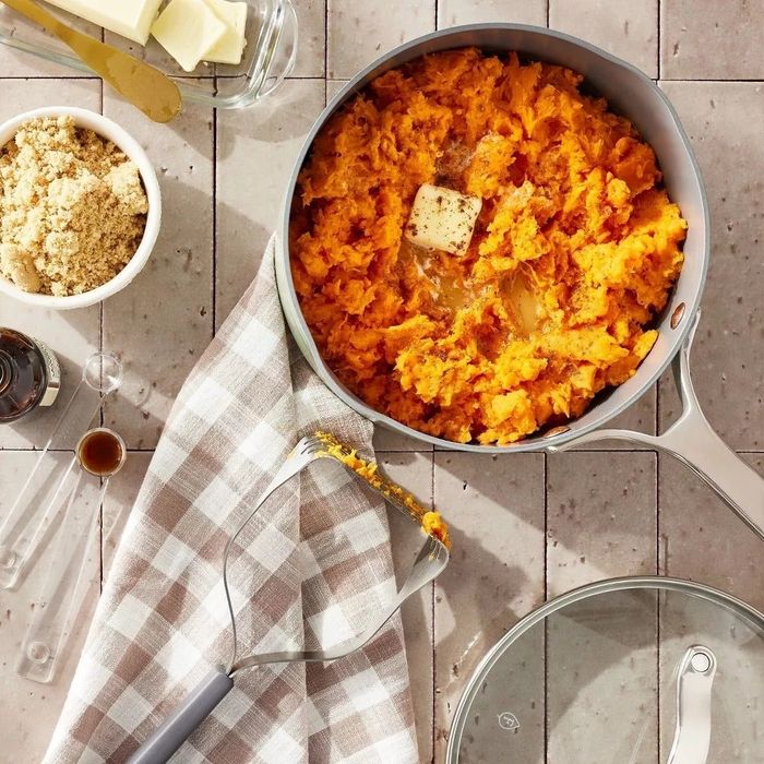 A pot filled with mashed sweet potatoes topped with a pat of butter, surrounded by kitchen essentials like brown sugar, vanilla extract, butter, measuring spoons, a towel, and a pot lid.