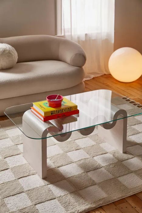 A glass coffee table with curved metal legs, adorned with books and a red bowl on top, placed in a modern living room with a plush sofa and a balloon-shaped floor lamp.