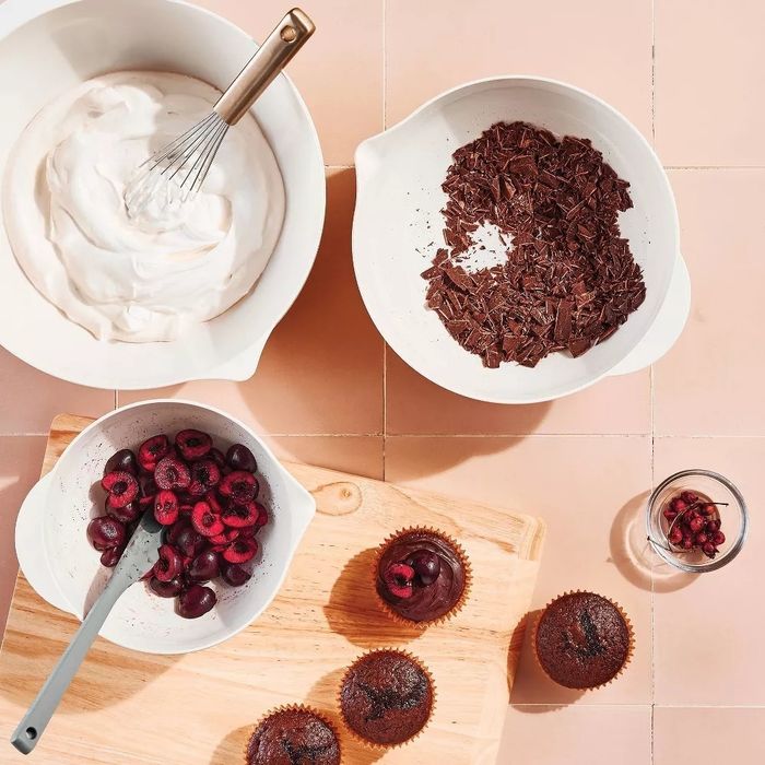 A baking setup featuring whipped cream, chocolate shavings, cherries, and chocolate cupcakes displayed on a wooden board, ideal for showcasing a dessert recipe.