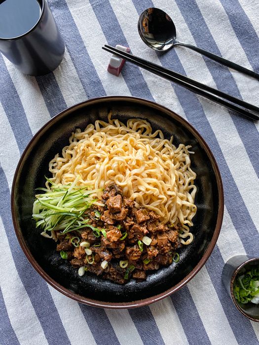 A bowl of noodles topped with diced meat, cucumber strips, and fresh green onions, accompanied by chopsticks and set on a striped tablecloth