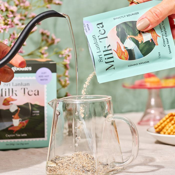 Hands pouring Sri Lankan milk tea mix and hot water into a glass mug on a table surrounded by snacks and flowers. Package in the background
