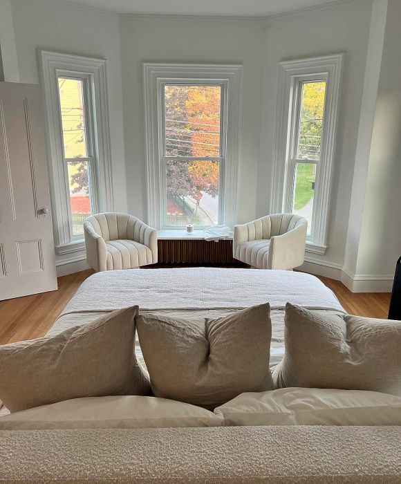 A cozy bedroom with a neatly arranged bed surrounded by plush pillows, two armchairs placed near three large windows, and a small table nestled between them.