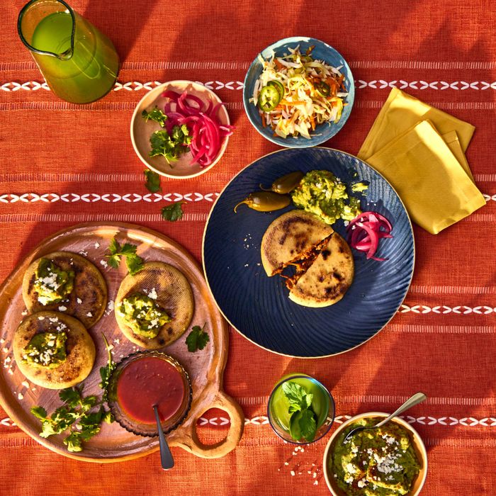 A colorful table setup featuring pupusas, salsa, pickled onions, and slaw served on decorative plates over a striped tablecloth