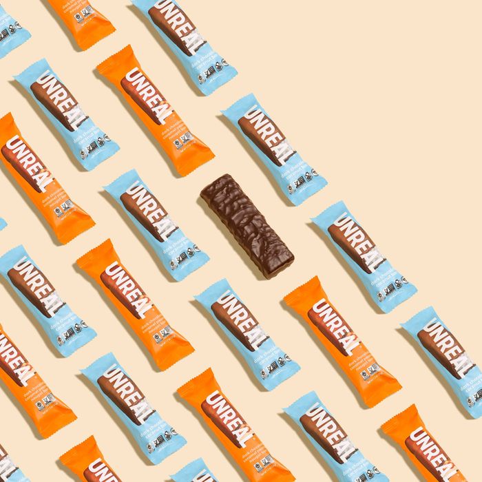 Unreal chocolate bars in vibrant orange and blue wrappers are arranged in a diagonal pattern, with one unwrapped bar placed in the center