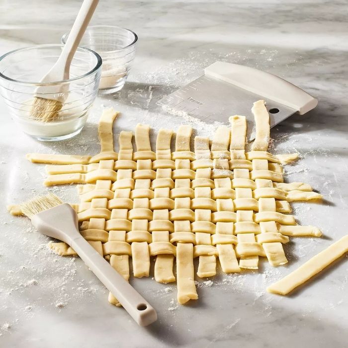 Pie dough is beautifully latticed on a floured marble surface, with two glass bowls, a pastry brush, and a bench scraper placed nearby.