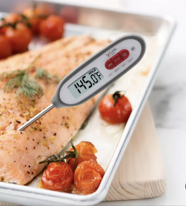 A Taylor digital waterproof instant-read thermometer, inserted into a piece of baked salmon, reads 145.3°F. Features include waterproofing, antimicrobial properties, and a hold function.