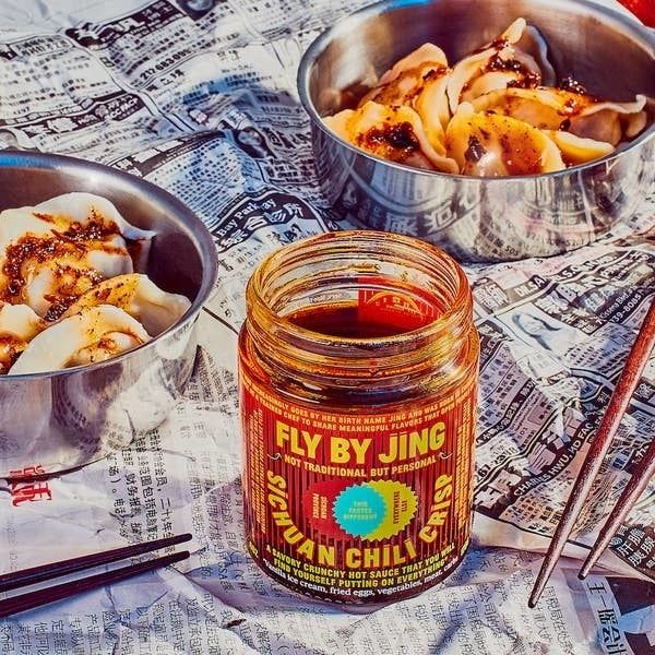 Fly By Jing's Sichuan Chili Crisp jar placed on a table beside two bowls of dumplings smothered in sauce, set against a newspaper backdrop with chopsticks on the side