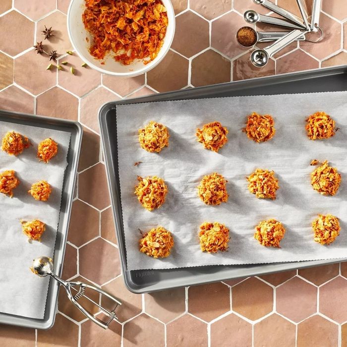 A baking sheet covered with scoops of carrot mixture on parchment paper, alongside a bowl of the mixture, measuring spoons, and a cookie scoop.