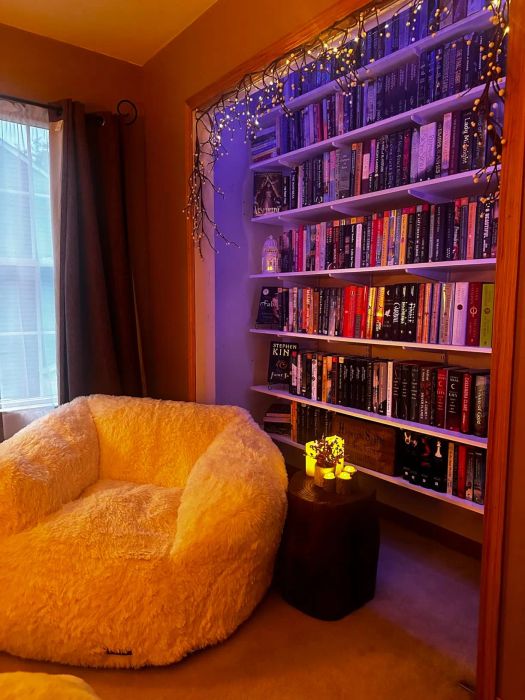 A cozy reading corner featuring a plush bean bag chair, a small side table with candles, and shelves brimming with books, all bathed in the warm glow of string lights.