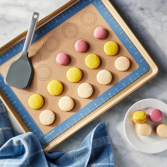 A baking tray featuring macarons in three different colors, with a spatula, a blue cloth beside it, and additional macarons resting on a white plate.