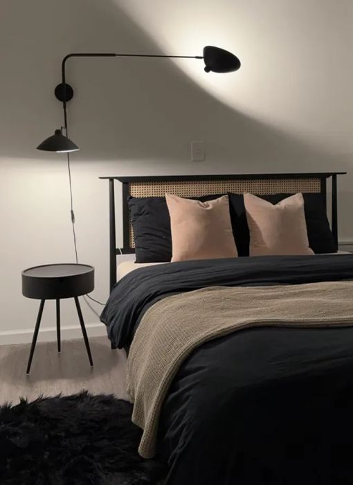 A modern bedroom featuring a black bed frame, beige pillows, a beige throw blanket, a black side table, a wall-mounted black lamp, and a soft black rug.