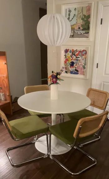 A round dining table with a white vase filled with vibrant flowers, surrounded by four chairs with green cushions and wooden backs. Modern art graces the background wall