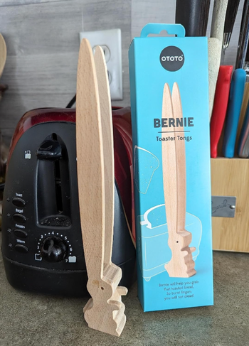 A pair of wooden toaster tongs designed like a cute bunny, displayed next to its packaging.