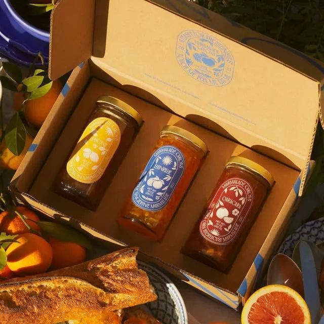 three jars of homemade marmalade in a cardboard box, surrounded by fresh bread and citrus fruits, set up for a picnic