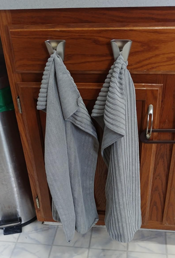Two gray kitchen towels draped over sleek metal hooks on a wooden cabinet door.