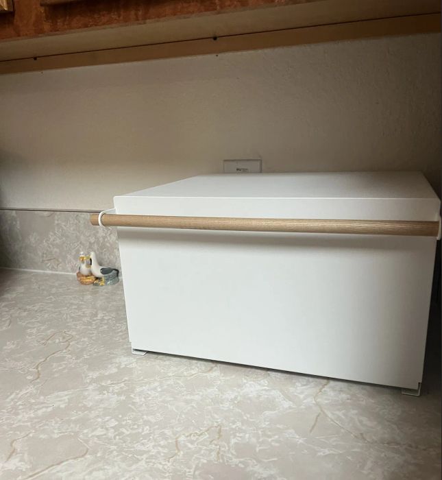 A modern white bread box with a wooden handle, sitting on a kitchen countertop under cabinets