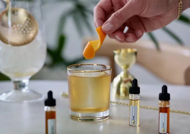 A hand garnishing a cocktail with a twist of orange peel. On the table, dropper bottles with various extracts and a luxurious gold jigger.