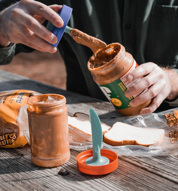 A person spreads peanut butter onto bread with a silicone spatula, surrounded by peanut butter jars on a picnic table