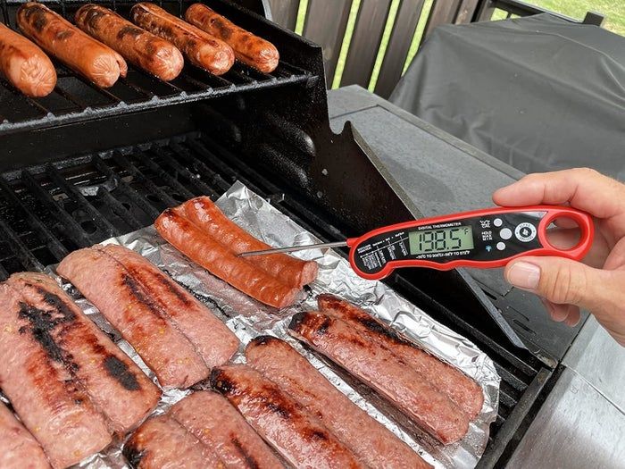 Grilling sausages and hot dogs, with a digital thermometer showing 188.5°F as it checks the temperature of one hot dog
