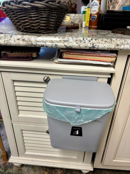 A small trash bin attached to a kitchen cabinet drawer, positioned under a counter alongside a basket and spices