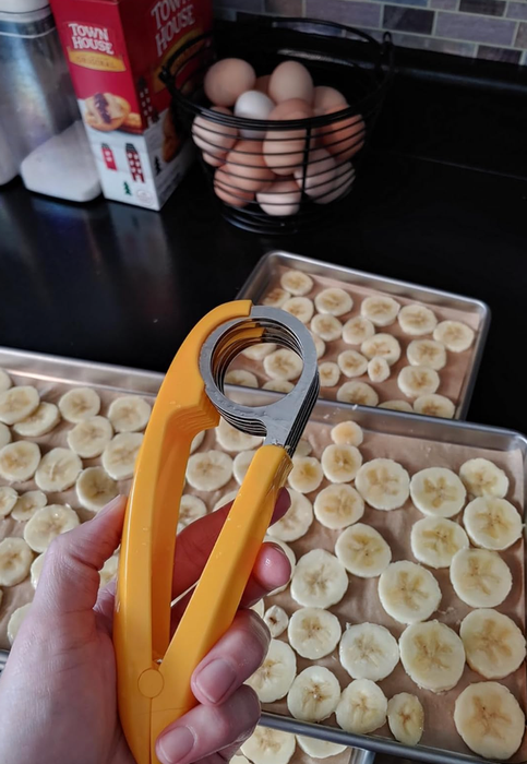 A hand holds a banana slicer above trays of neatly sliced bananas, with eggs and crackers visible on the counter