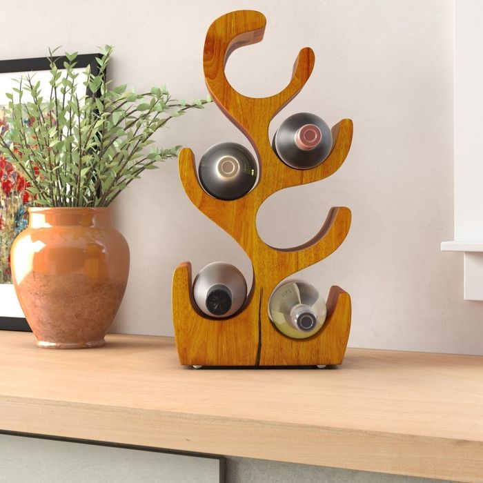 A wooden wine rack holding four bottles, placed on a countertop next to a vase with green leaves and a framed picture in the background.