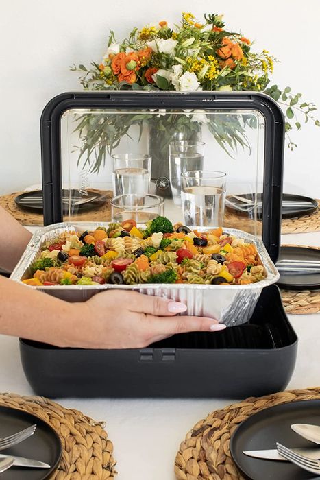 A hand placing a foil pan of pasta salad into the sturdy black protector, with its tall plastic sides for extra support.