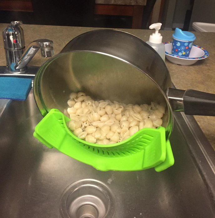 A reviewer's photo showing the green strainer clipped to the side of a pot, successfully straining the water while keeping the macaroni shells inside.