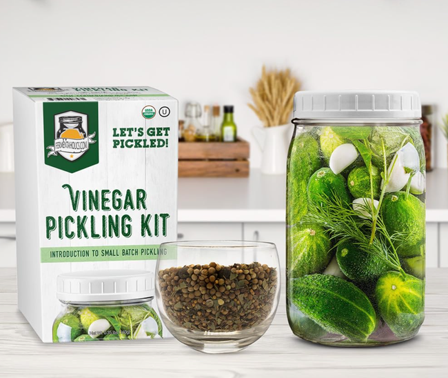 A vinegar-based pickling kit displayed alongside a jar of pickled vegetables and a bowl of spices on a kitchen countertop.