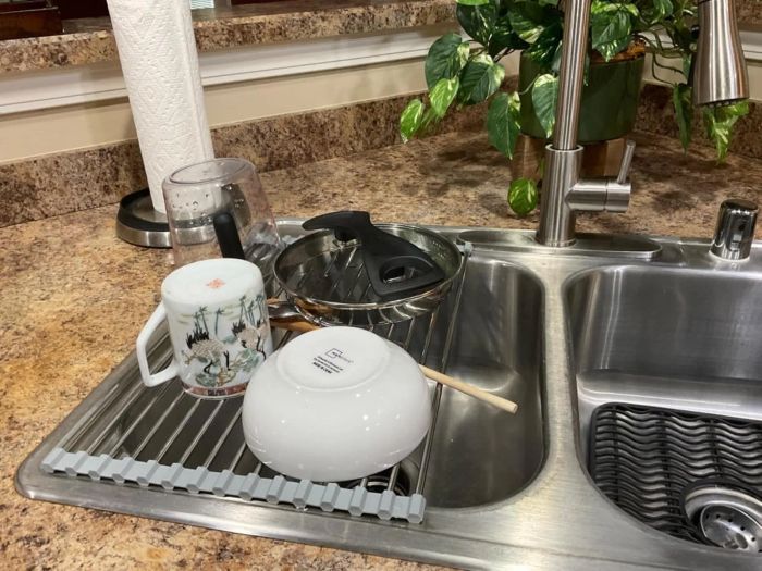A kitchen sink with dishes drying on a rack, including a mug, bowl, pot lid, and utensils, with a plant and a paper towel roll in the background