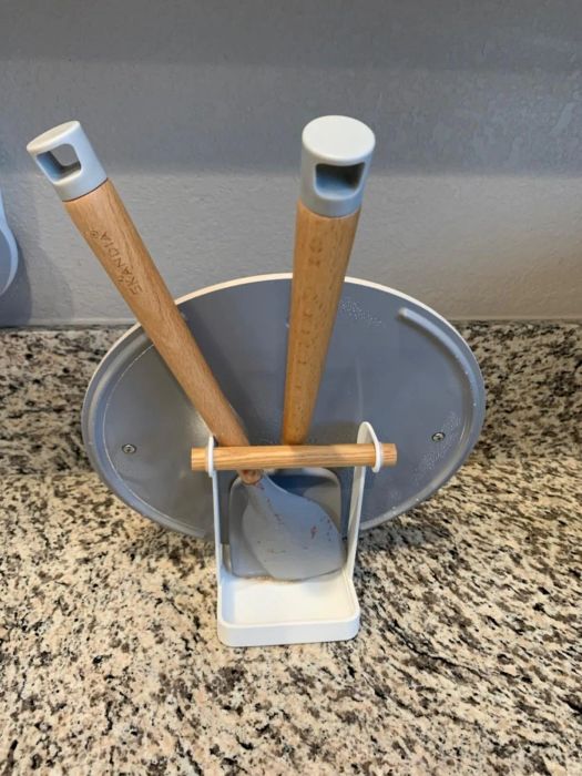 A kitchen utensil and lid holder on a granite countertop, holding a pan lid alongside a wooden-handled spatula and spoon