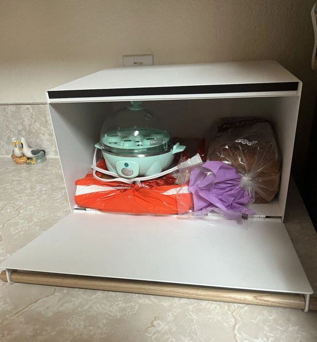 A bread box containing bread, an egg cooker, and a bag tucked behind a flap