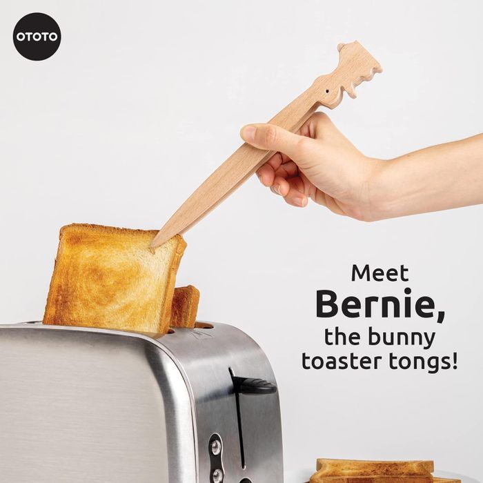 A hand using Bernie, the wooden bunny-shaped toaster tongs, to safely retrieve toast from a toaster. Product by OTOTO.