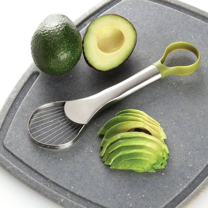 An avocado slicer placed next to a halved avocado and slices neatly arranged on a cutting board, demonstrating its use for a Shopping category article.