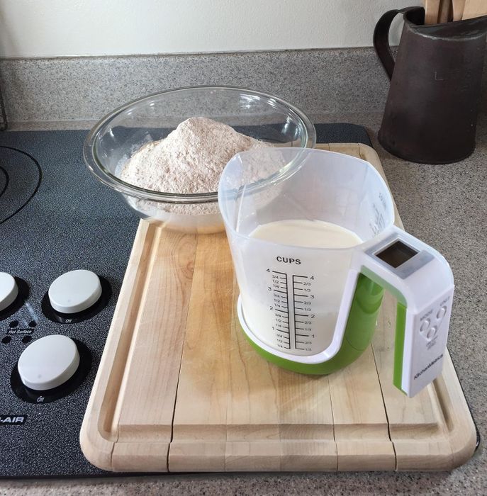 A bowl of flour and a measuring cup of milk resting on a wooden cutting board next to a stove.
