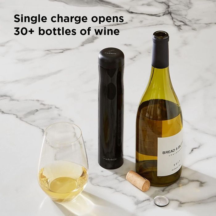An electric wine opener alongside a wine bottle, a glass filled with wine, a cork, and its wrapper, all arranged on a marble surface. Text: 