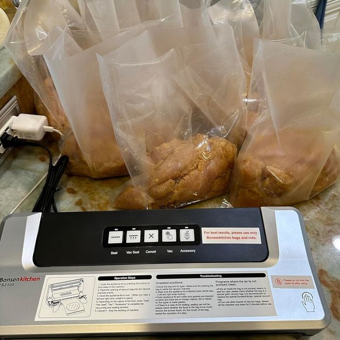 A vacuum sealing machine sits on a counter, surrounded by multiple vacuum-sealed bags of food, with clear instructions for proper use displayed on the machine.