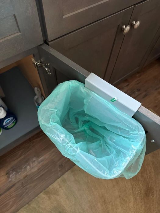 A small trash bin with a green liner attached to the inside of a cabinet door. Cleaning supplies are visible inside the cabinet