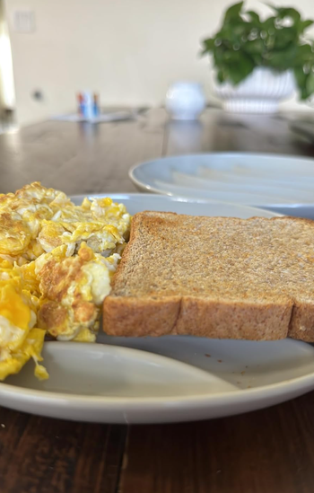 Scrambled eggs and toast served on a white plate, resting on a wooden table with a plant softly blurred in the background