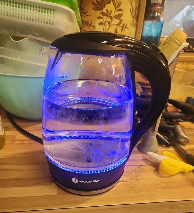 A sleek electric glass kettle filled with water, illuminated by blue LED lights, placed on a wooden countertop surrounded by various kitchen utensils.