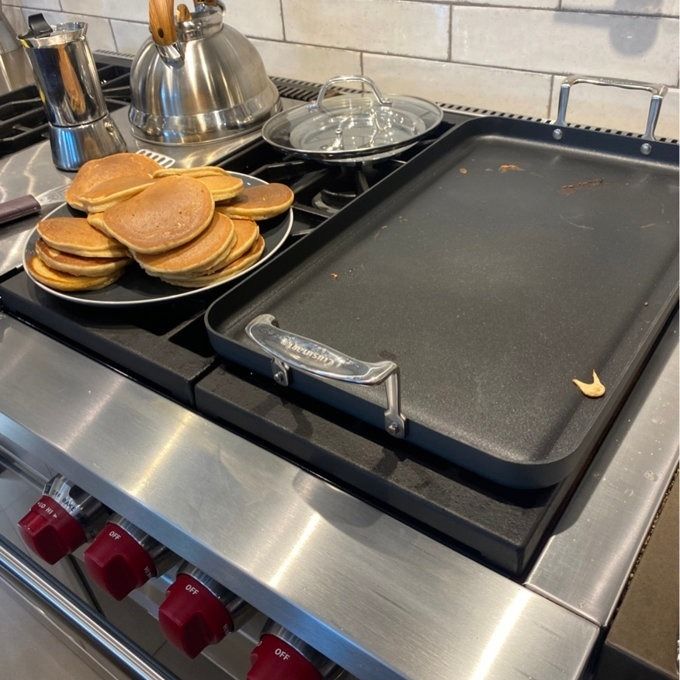 A large stove with a griddle, a plate of stacked pancakes, a kettle, and a coffee maker placed beside it.