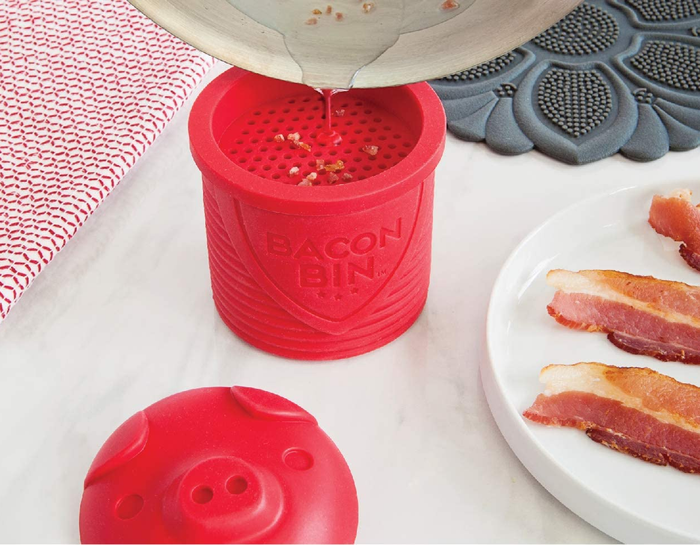 Bacon grease being poured into the red bacon bin, placed next to a plate of crispy bacon