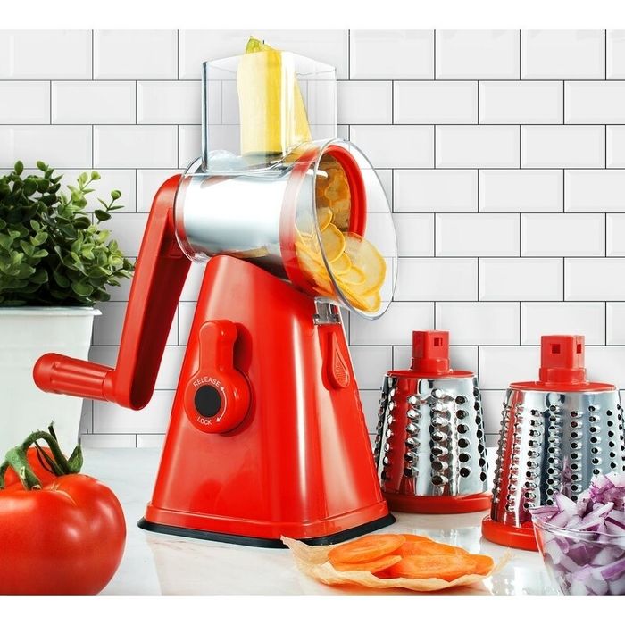 A manual vegetable slicer on a kitchen counter, with slices of vegetables, a tomato, and two different shredding attachments displayed nearby.
