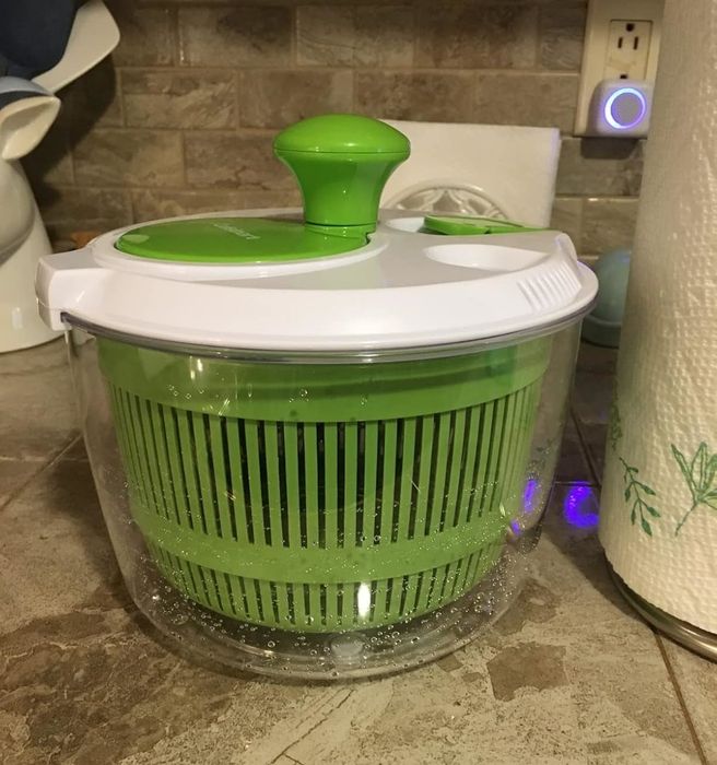 A plastic salad spinner sitting on a kitchen countertop, with a green handle and white lid. Inside, there’s a green inner basket placed within a clear plastic bowl.