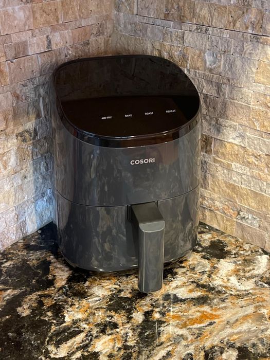 A Cosori air fryer sits on a marble countertop with a stone backsplash. It features buttons for air frying, baking, roasting, and reheating.