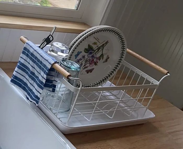 A kitchen sink with a window view, featuring a dish rack holding plates, a glass jar, and a striped towel, with trees visible outside the window