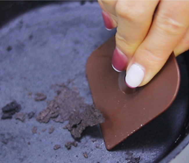 A hand scrapes leftover residue from a cast iron skillet using a brown scraping tool.