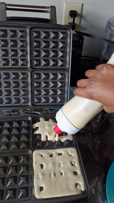 A hand pouring batter into a waffle iron.