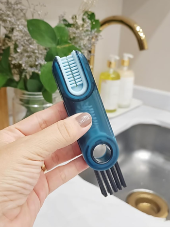 A hand holding a kitchen brush cleaner with bristles in front of a sink and soap dispensers
